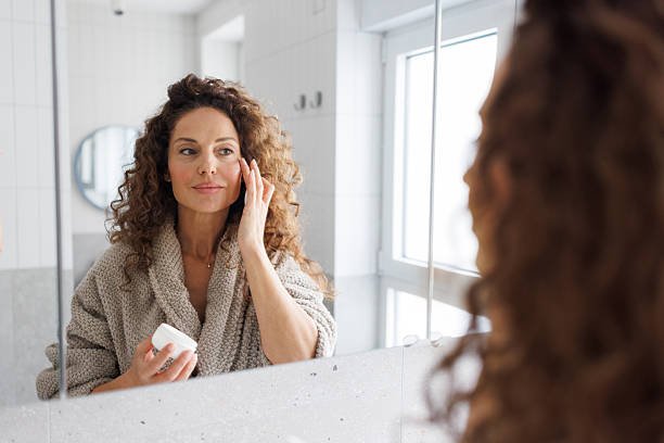 A mid adult woman with curly hair wearing a cozy bathrobe doing skiincare face cream to her face in a bright, modern bathroom with natural daylight streaming through the window.