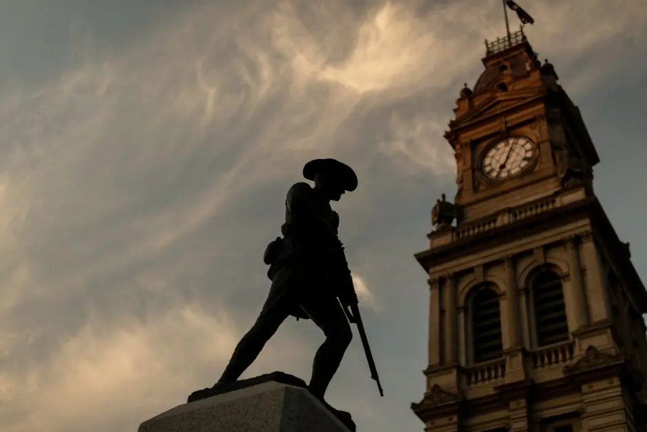 Silhouette of a war memorial statue against a dramatic sky with a historic clock tower in the background at sunset.